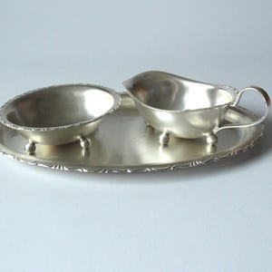 May include: A silver-toned serving set featuring a small bowl, a gravy boat with a handle, and an oval tray. The bowl and gravy boat rest on small, rounded feet. The tray has a decorative, scalloped edge, and the set is likely vintage.