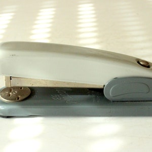 Puede incluir: Una grapadora de metal gris con la parte superior blanca. La grapadora tiene un removedor de grapas plateado y las palabras "Swingline 53" impresas en el lateral.