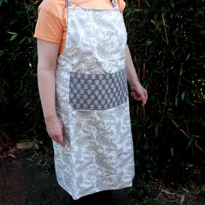 May include: A white apron with a paisley pattern and a gray pocket. The apron has adjustable straps.