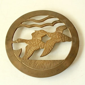 Könnte beinhalten: Ein runder, polierter Messing-Topfuntersetzer mit einem Design aus zwei fliegenden Enten und Wellenlinien.