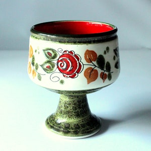 Op de afbeelding: Een groene en witte keramische voetkom met een rode binnenkant en een bloemenontwerp met rode rozen, groene bladeren en oranje accenten.