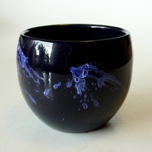Pode incluir: Uma tigela de cerâmica azul escura com um esmalte branco salpicado. A tigela tem uma forma arredondada e um acabamento liso e brilhante.