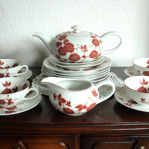 Könnte beinhalten: Ein weißes Porzellan-Teeservice mit rotem Blumenmuster. Das Set beinhaltet eine Teekanne, einen Sahnekännchen, Untertassen und Teetassen. Das Teeservice wird auf einer dunklen Holzoberfläche präsentiert.