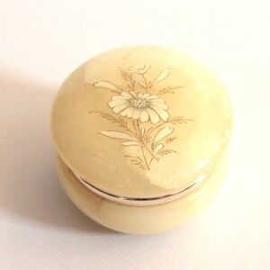 May include: A round, cream-colored alabaster trinket box with a floral design. The lid features a delicate white flower with a gold outline and the word "Flora" inscribed on the bottom right.