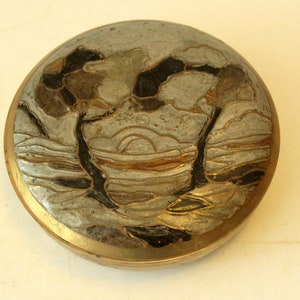 May include: A round brass trinket box with a silver and black design of trees and a river. The box has a raised rim and a smooth surface.