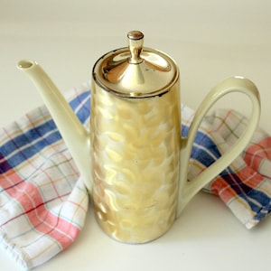 Può includere: Una caffettiera in ceramica dorata e bianca con un design testurizzato e un coperchio. La caffettiera è appoggiata su un tovagliolo a quadri.