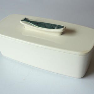 Pode incluir: Prato retangular de cerâmica creme com tampa. A tampa apresenta uma pega em relevo com um desenho de peixe verde-azulado. O prato é provavelmente para servir ou armazenar alimentos.