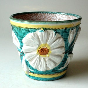 May include: A ceramic planter with a turquoise base and a large white daisy design. The planter has a yellow and gold trim, and the interior is speckled with brown and white. The planter is approximately 15 cm tall.
