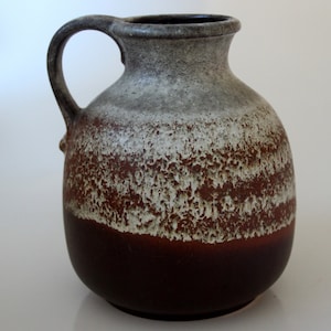 Könnte beinhalten: Eine braune und weiße Keramikvase mit texturierter Glasur. Die Vase hat einen breiten, runden Körper und einen schmalen Hals mit einem Henkel.