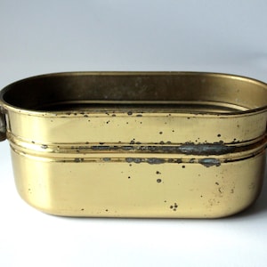 Peut inclure: Un récipient rectangulaire en laiton avec des coins arrondis et deux petites poignées. Le récipient a un aspect terni et vieilli avec des taches sombres et une surface légèrement inégale. L'intérieur est visible et semble vide.