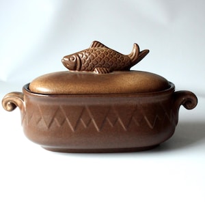 Op de afbeelding: Bruine keramische ovenschaal met een deksel en een visvormige handgreep. De schaal heeft een getextureerd oppervlak en een decoratief zigzagpatroon rond de basis. De handvatten zijn gebogen.