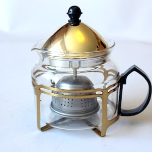 Könnte beinhalten: Eine Teekanne aus klarem Glas mit einem glänzenden, goldfarbenen Metallrahmen und Boden. Der Deckel ist mit einem kleinen schwarzen Knopf versehen und der Griff besteht aus schwarzem Kunststoff. Im Inneren befindet sich ein Teesieb aus Edelstahl.