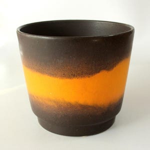 Könnte beinhalten: Keramik-Blumentopf mit dunkelbraunem Ober- und Unterteil sowie einem leuchtend orangefarbenen Band in der Mitte. Der Topf hat eine zylindrische Form und eine leicht strukturierte Oberfläche, geeignet für den Innen- und Außenbereich.