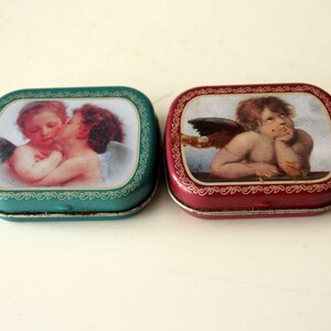 May include: Two small, square, metal tins with images of cherubs. One tin is teal with gold trim and features two cherubs embracing. The other tin is red with gold trim and features a single cherub with wings, looking thoughtful.