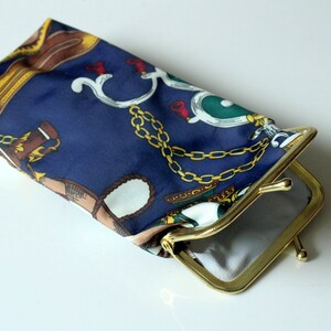 Puede incluir: Una cartera de monedas de tela azul con un marco de metal dorado y un diseño de cadena. La cartera está abierta y el interior está forrado con tela blanca.