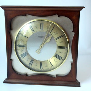 Könnte beinhalten: Eine Uhr mit Holzrahmen, goldenem Zifferblatt und römischen Ziffern. Das Zifferblatt der Uhr zeigt "Dugena Quartz".