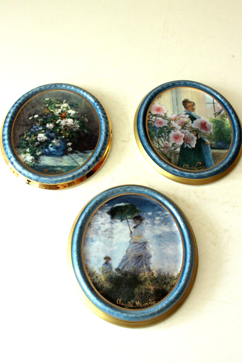 Pode incluir: Tr&ecirc;s pratos redondos de cer&acirc;mica com acabamento dourado e bordas azuis. Cada prato apresenta uma pintura diferente: um buqu&ecirc; de flores, uma mulher segurando um buqu&ecirc; de flores e uma mulher com um guarda-chuva caminhando em um campo.