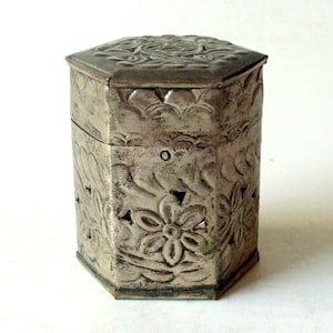 Pode incluir: Uma caixa hexagonal de prata com intrincados desenhos florais em relevo na superfície. A caixa tem uma tampa e é feita de metal.