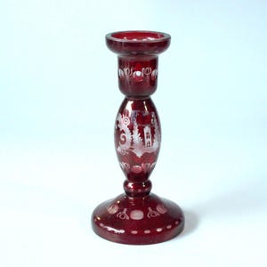 May include: A ruby red glass candlestick with etched white designs. The candlestick features a round base, a bulbous center section with a detailed landscape scene, and a top cup for holding a candle. The overall height is approximately 18 cm.