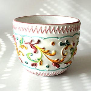 Könnte beinhalten: Ein weißer Keramikübertopf mit einem erhabenen, handbemalten Reliefdesign aus floralen Wirbelmustern in Rot, Gelb und Grün. Der Rand und der Boden sind mit einem roten Zickzackmuster verziert und die Oberfläche weist eine Textur auf.
