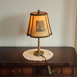 Könnte beinhalten: Eine Vintage-Tischlampe mit beigem Lampenschirm mit einer gerahmten pastoralen Szene. Die Lampe hat einen Messingfuß und ein dunkelbraunes Kabel. Sie steht auf einem dunklen Holztisch mit einer weißen Spitzendecke.