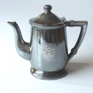 Op de afbeelding: Een zilverkleurige theepot met een deksel en een gebogen tuit. De theepot heeft een handvat en een klein embleem op de voorkant. De theepot is waarschijnlijk van metaal en staat op een wit oppervlak. Het is een vintage item.