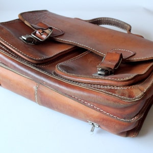 May include: A brown leather briefcase featuring two front pockets with metal buckle closures, a top handle, and visible white stitching along the edges. The bag has a structured rectangular shape with metallic hardware attachments on the sides.