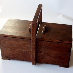 Op de afbeelding: Een vintage houten naaidoos met een handvat. De doos is rechthoekig met een scharnierend deksel en een klein houten handvat aan de bovenkant. Het hout heeft een rijke, donkerbruine kleur en zichtbare nerf.