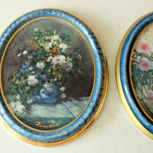 Pode incluir: Dois pratos redondos de cer&acirc;mica com guarni&ccedil;&atilde;o dourada e bordas azuis. Os pratos apresentam imagens emolduradas de arranjos florais. O prato da esquerda apresenta um buqu&ecirc; de flores em um vaso azul com a assinatura do artista "Renoir" em letras douradas. O prato da direita apresenta um arranjo floral com a assinatura do artista "Chardin" em letras douradas.