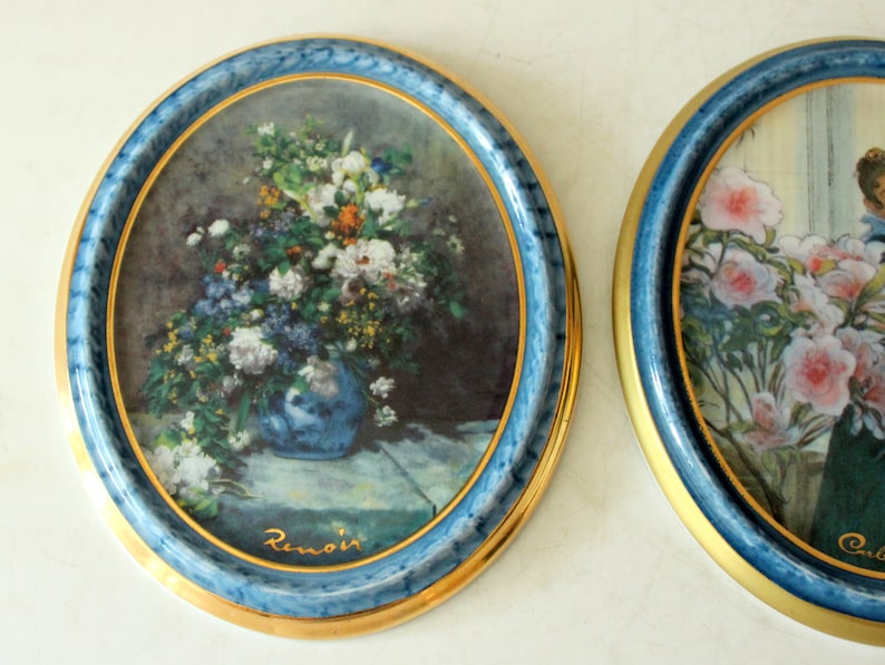 Pode incluir: Dois pratos redondos de cer&acirc;mica com guarni&ccedil;&atilde;o dourada e bordas azuis. Os pratos apresentam imagens emolduradas de arranjos florais. O prato da esquerda apresenta um buqu&ecirc; de flores em um vaso azul com a assinatura do artista "Renoir" em letras douradas. O prato da direita apresenta um arranjo floral com a assinatura do artista "Chardin" em letras douradas.