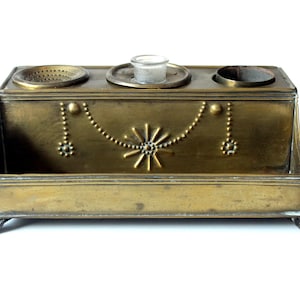 May include: Antique brass desk organizer with three compartments and a small glass insert. The rectangular box features decorative beaded accents and a starburst design. The organizer sits on small, curved feet.