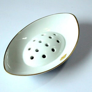 May include: White ceramic flower frog with a gold rim and 12 holes. The frog is oval shaped and sits on a white surface.