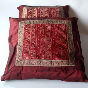 Op de afbeelding: Twee bordeauxrode decoratieve kussens. Het grotere kussen heeft een vierkant, geborduurd midden met een gouden bloemenontwerp. Het kleinere kussen heeft een vergelijkbaar ontwerp. De kussens zijn gemaakt van een glanzend, zijdeachtig materiaal.