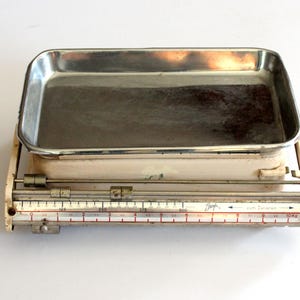 Op de afbeelding: Vintage crèmekleurige weegschaal met een roestvrijstalen bak. De weegschaal heeft een schuifbare gewichtsindicator met rode en zwarte markeringen, die tot 10 kg meet. De wijzerplaat heeft de tekst "zum tarieren".