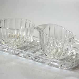 Könnte beinhalten: Ein Set aus Zucker- und Sahneschüssel sowie Serviertablett aus klarem, geschliffenem Glas. Schüssel und Sahnekännchen haben vertikale Rillen, das Tablett ein Gittermuster. Das Set ist zum Servieren von Zucker und Sahne bestimmt.