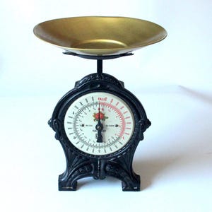 May include: A black cast iron kitchen scale with a gold-toned metal bowl on top. The circular white dial features black numerical markings, red measurement indicators, and a central floral graphic with a red rose and green leaves.