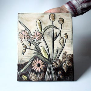 May include: A rectangular ceramic tile featuring a raised relief of stylized flowers and foliage. The flowers are a soft pink, with green stems and leaves. The background is a light beige with a wood grain texture. The tile is held by a person.