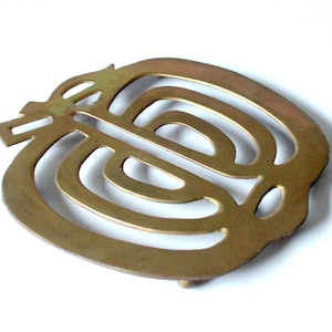 Könnte beinhalten: Ein messingfarbener Metall-Topfuntersetzer mit dekorativem, durchbrochenem Design. Der Topfuntersetzer weist eine Reihe von geschwungenen Linien und Formen auf, die ein abstraktes Muster bilden. Er hat kleine Füße.