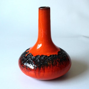 Op de afbeelding: Een kleine, oranje keramische vaas met een zwarte glazuur rond de bodem. De vaas heeft een rond lichaam en een lange, dunne hals.