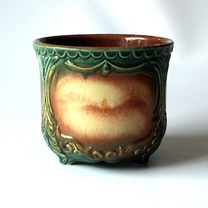 Può includere: Un vaso decorativo in ceramica di forma rettangolare. Il vaso presenta un esterno verde e marrone con un pannello centrale nei toni del giallo e del rosso. Il bordo e la base hanno un design smerlato.