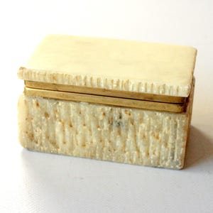 May include: A rectangular, off-white trinket box with a textured surface and a hinged lid. The lid and base are connected by two thin, gold-colored bands. The box has a rough, natural appearance, suggesting it may be made of stone or a similar material.