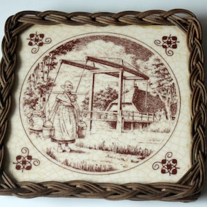 Puede incluir: Un azulejo de cerámica marrón y blanco con una imagen marrón y blanca de una mujer con un vestido tradicional holandés de pie frente a un puente. El azulejo está enmarcado en un marco de mimbre tejido.
