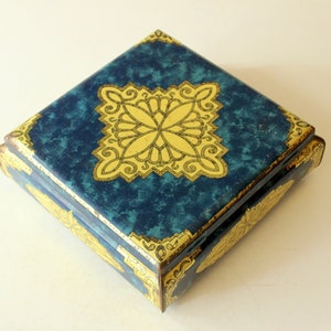 Op de afbeelding: Vierkante, vintage sieradendoos met een blauwe gemarmerde afwerking en gouden accenten. Het deksel heeft een centraal gouden bloemmotief en gouden hoekdetails. De doos is waarschijnlijk van metaal.