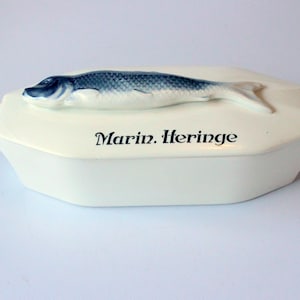 May include: A white ceramic serving dish with a blue and white fish-shaped lid. The dish is labeled "Marin. Heringe" in black text. The fish has detailed scales and fins.