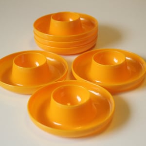 Peut inclure: Un ensemble de six coquetiers orange vif. Chaque coquetier est composé d'une base circulaire avec une partie centrale surélevée pour maintenir l'œuf. Les coquetiers sont en plastique brillant et sont disposés sur une surface blanche.