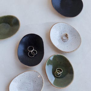 May include: A collection of small, oval-shaped ceramic ring dishes in various colors: black, white, and green. Each dish has a speckled finish and a raised edge. Some dishes hold gold rings.