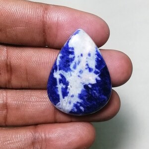 May include: A teardrop-shaped cabochon gemstone with a blue and white marbled pattern. The stone is a deep blue with white veins and swirls throughout.