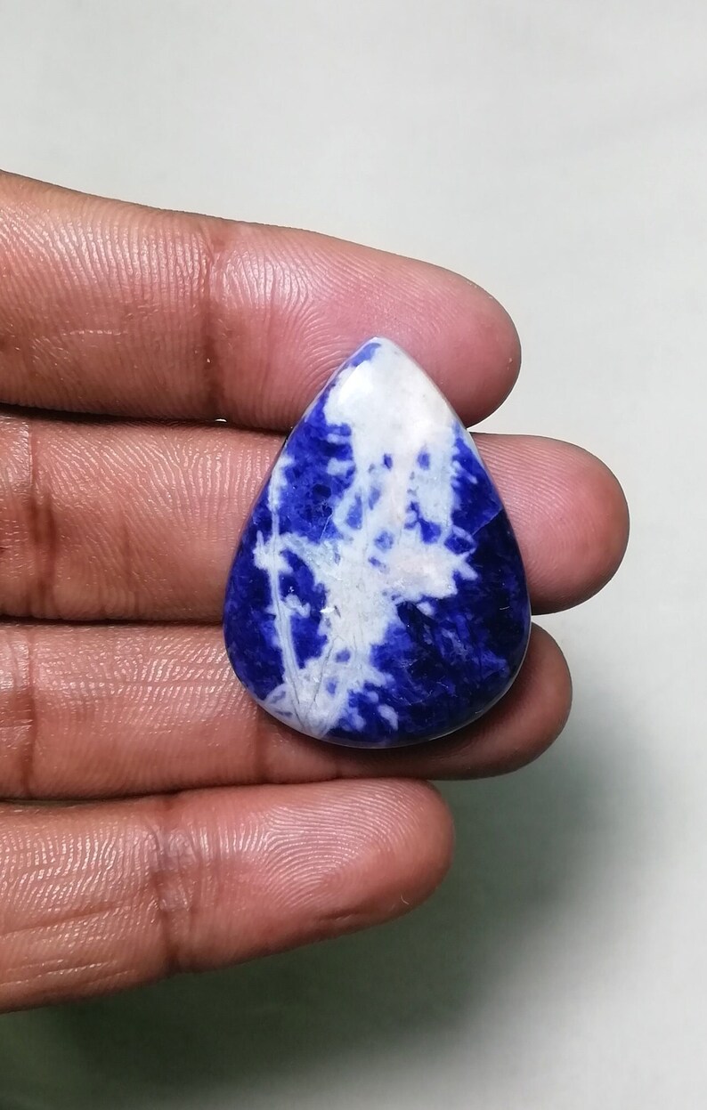 May include: A teardrop-shaped cabochon gemstone with a blue and white marbled pattern. The stone is a deep blue with white veins and swirls throughout.