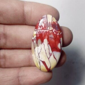 Może przedstawiać: Owalny kamień szlachetny w szlifie kaboszonowym z czerwonymi, białymi i żółtymi wzorami w kształcie wirów.