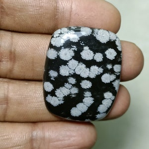 Puede incluir: Una piedra de obsidiana copo de nieve pulida y rectangular. La piedra es negra con patrones blancos en forma de copo de nieve. La piedra se sostiene en una mano, mostrando sus bordes lisos y redondeados y su diseño natural único.
